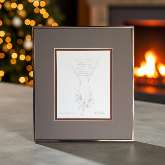 Framed artwork of holding hands on a table with a blurred Christmas tree and fireplace in the background