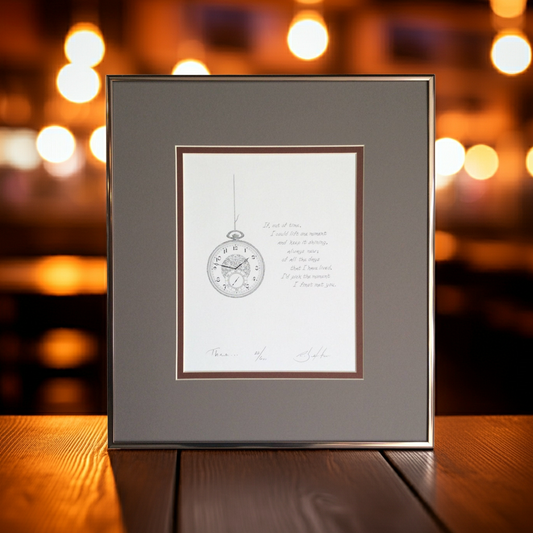 Framed artwork with a clock illustration on a wooden surface with blurred lights in the background